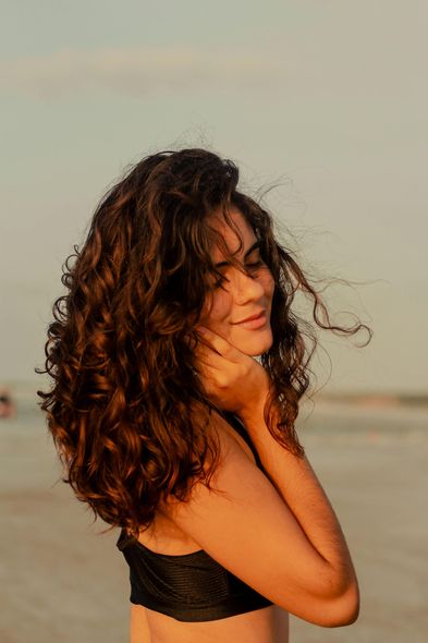 A woman with curly hair is standing on the beach with her hand on her face.