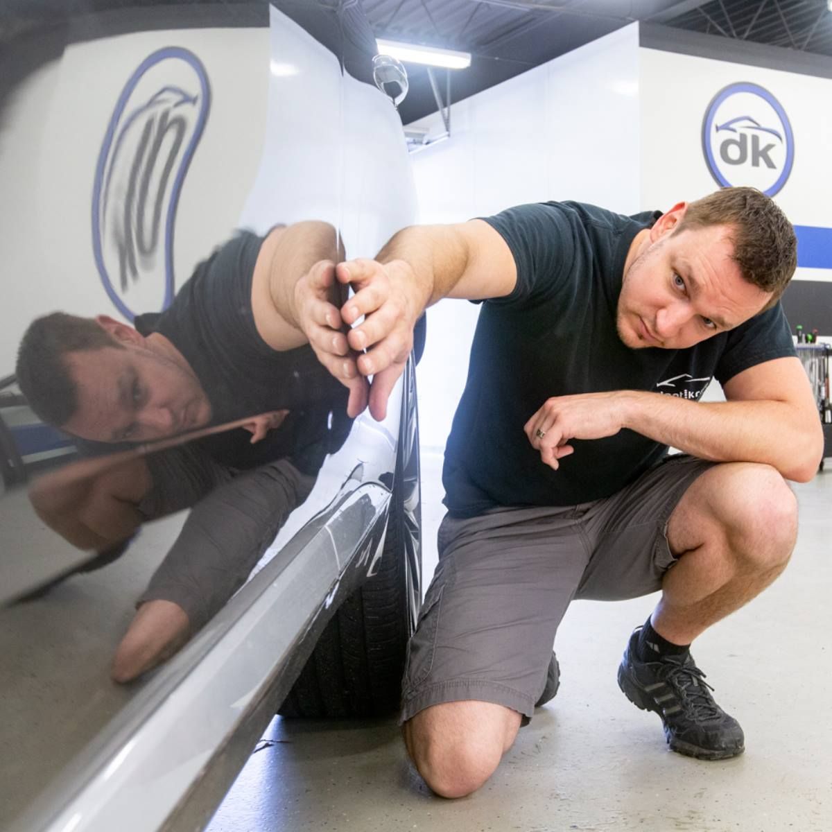 A man is kneeling next to a car with a dk logo on the wall behind him