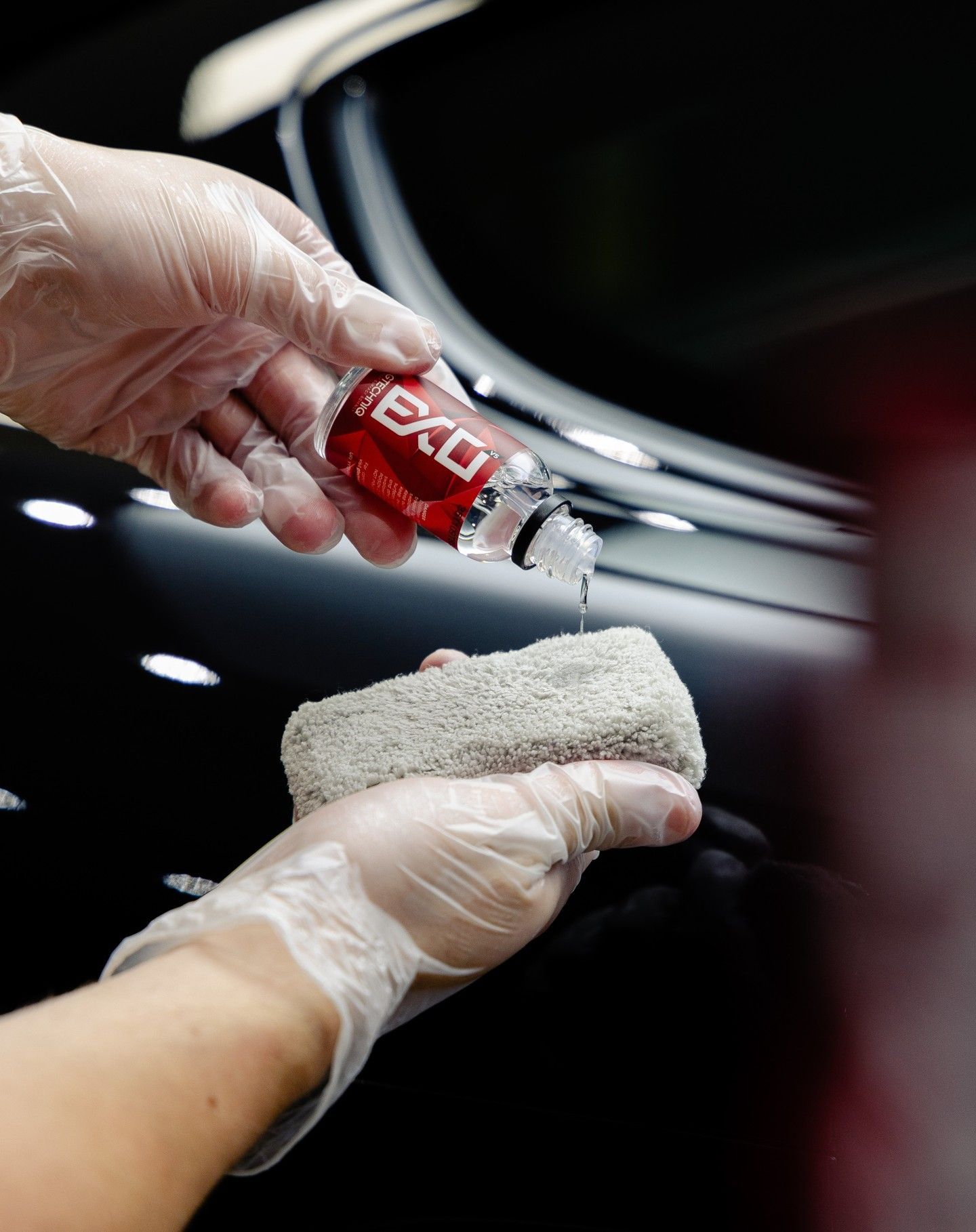 Gloved hands applying a liquid sealant from a red bottle onto a gray applicator pad on a black car surface.