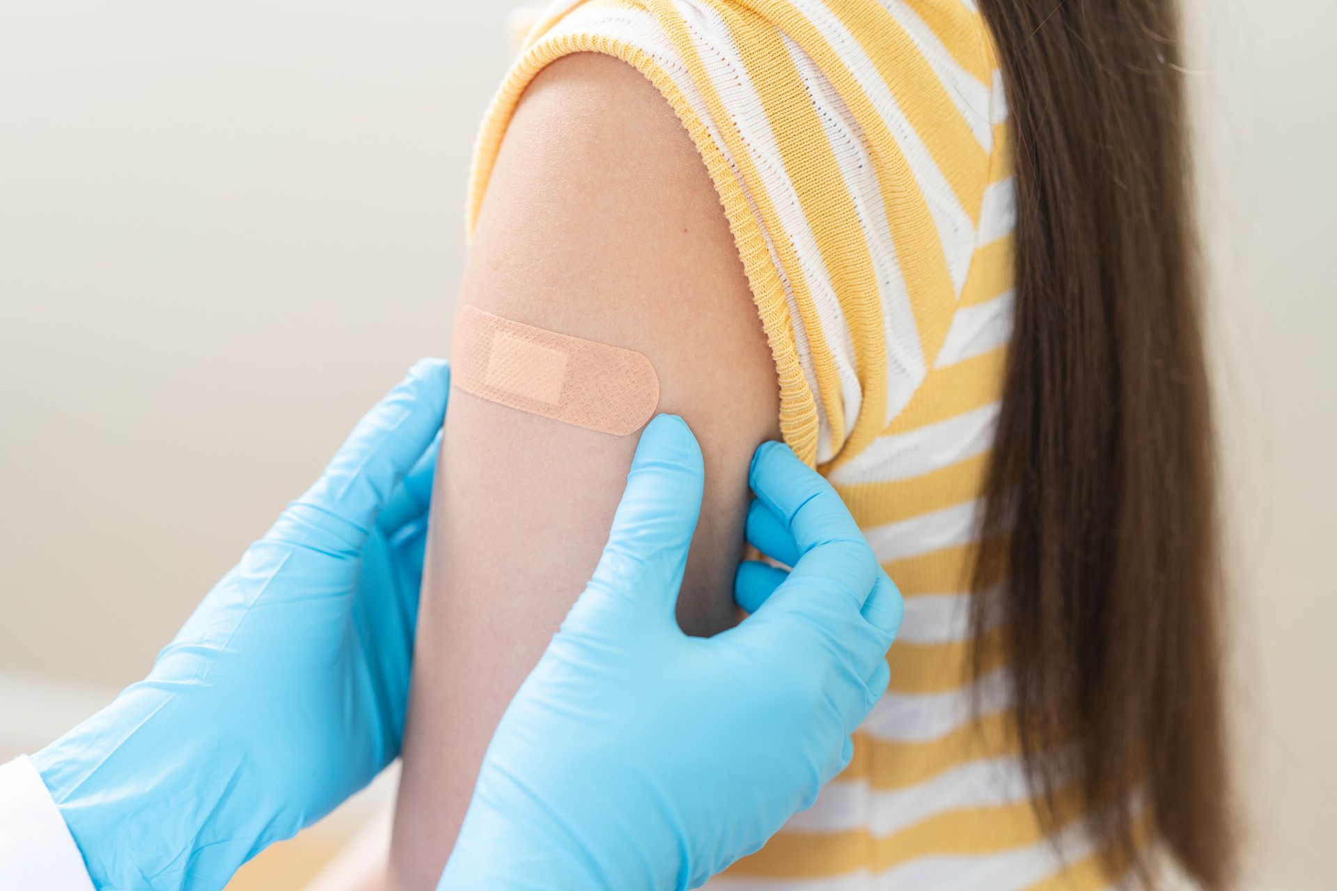 A girl with a bandage in her upper arm after receiving a vaccination. Gloved hands hold her arm.