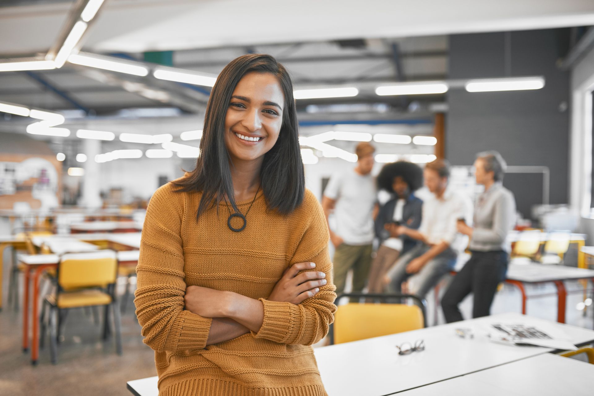 A woman smiling at the camera with her arms crossed in an office with colleagues in the background.