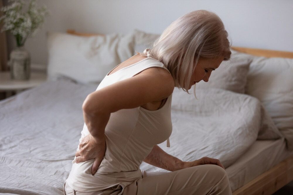 A Woman is Sitting on a Bed Holding Her Back in Pain — Life24 Health Bowen Therapy In Lavington, NSW
