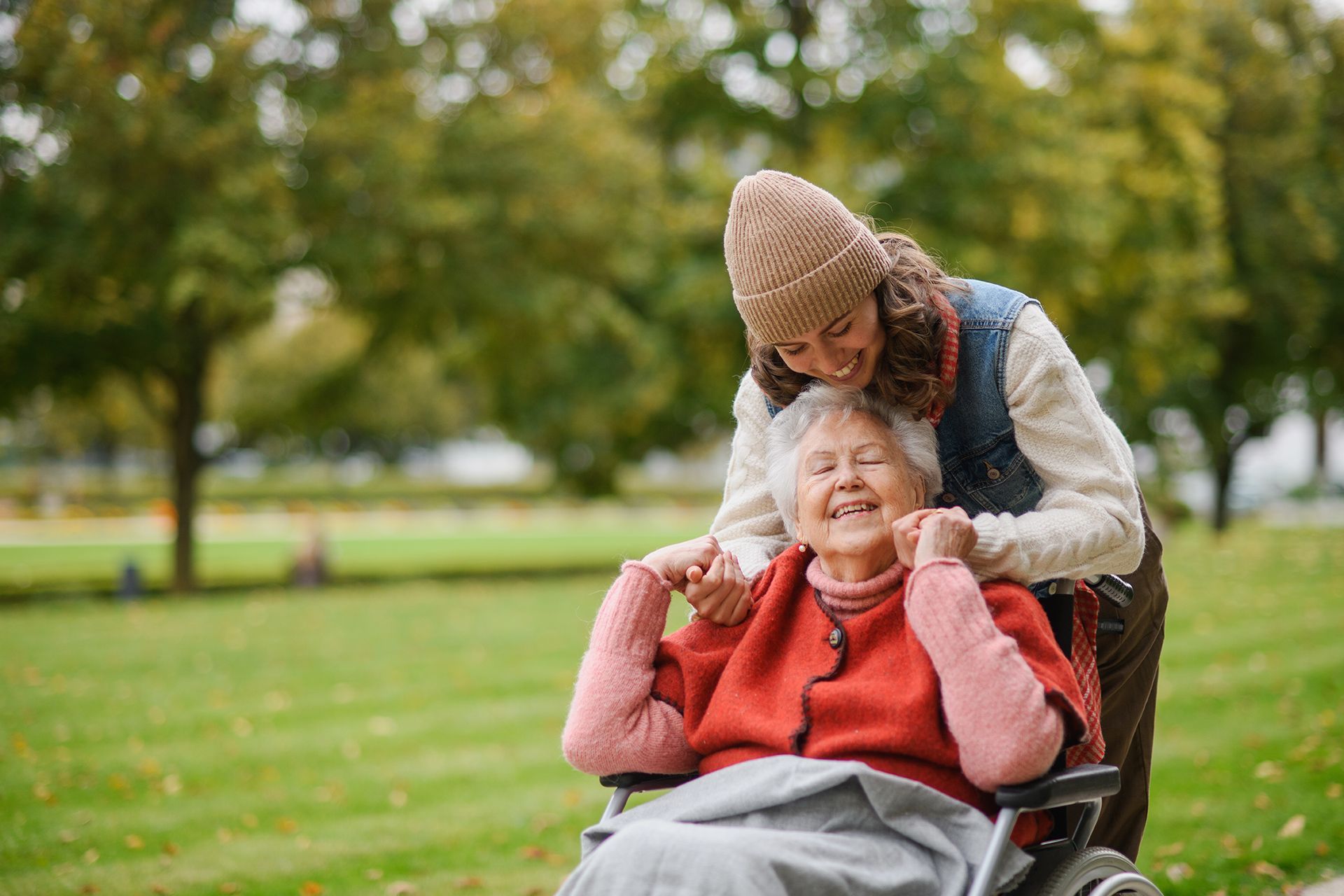 Woman assists elderly person in wheelchair outdoors; both smiling.