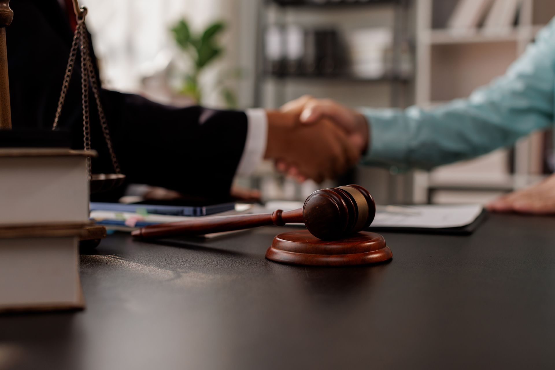 Gavel on desk with legal books; blurred view of two people shaking hands, likely a legal agreement.