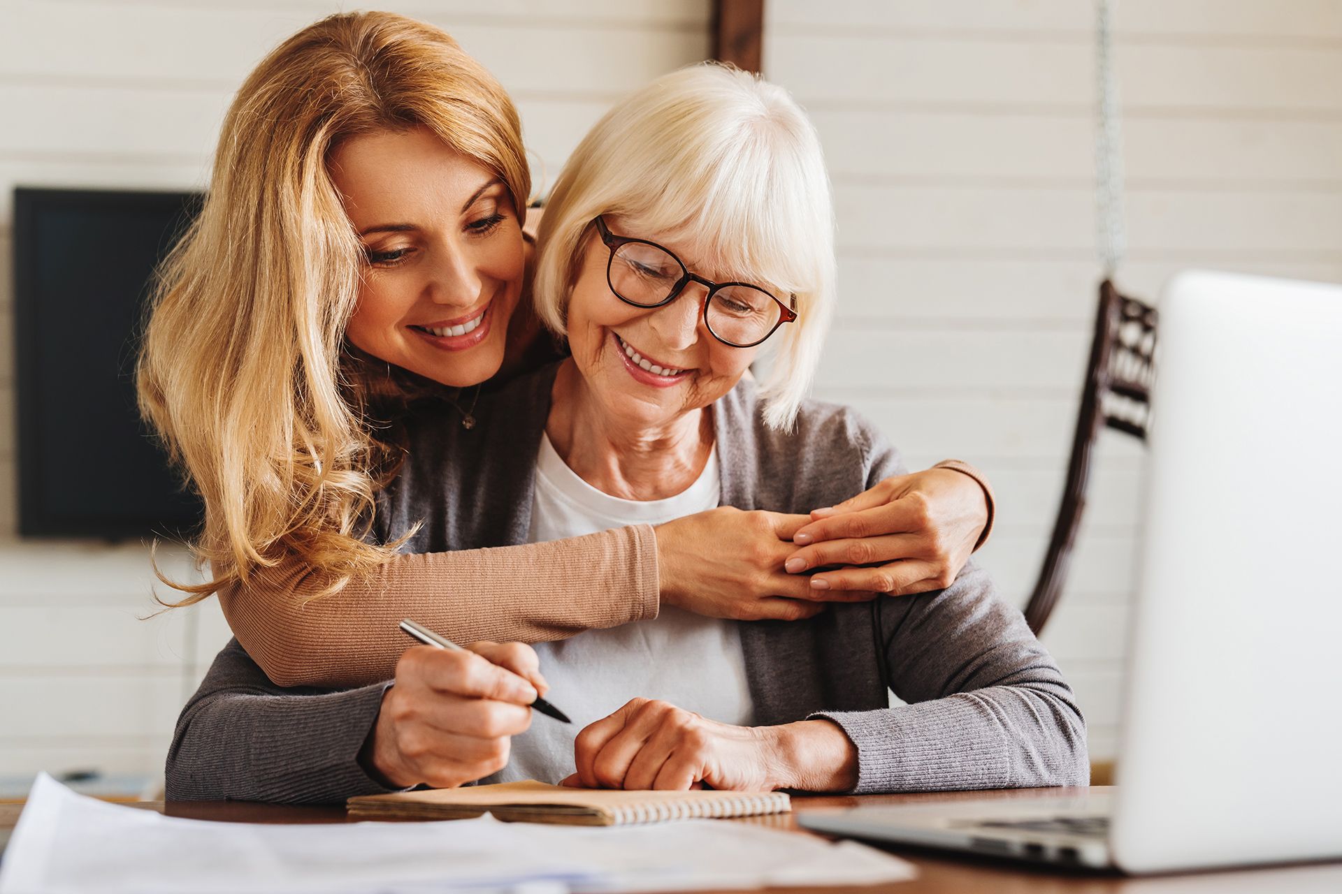 Woman hugs another woman, helping her write at a table with a laptop. Both are smiling.