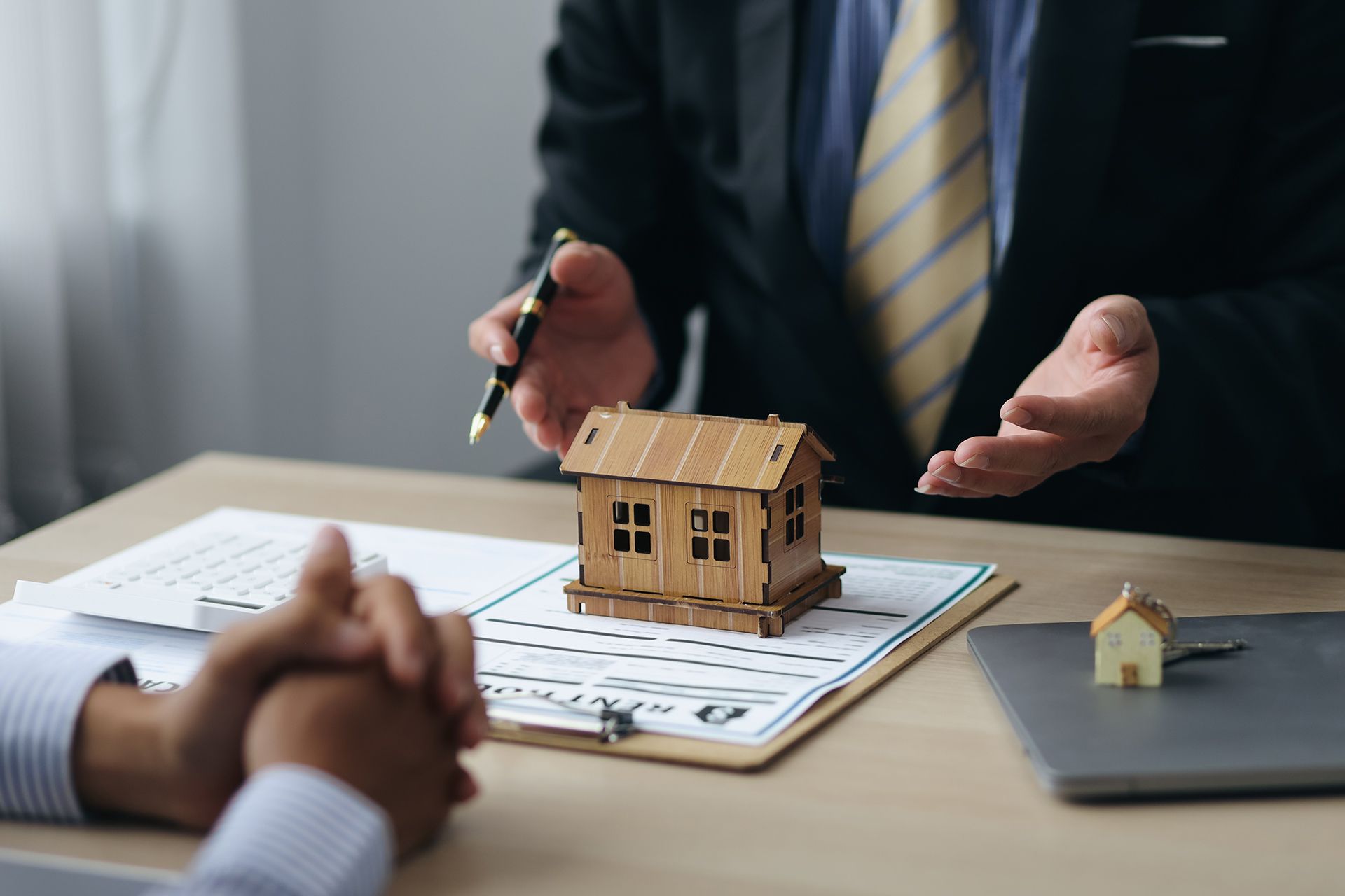 Real estate agent gesturing, discussing paperwork with a client, wooden house model on desk.