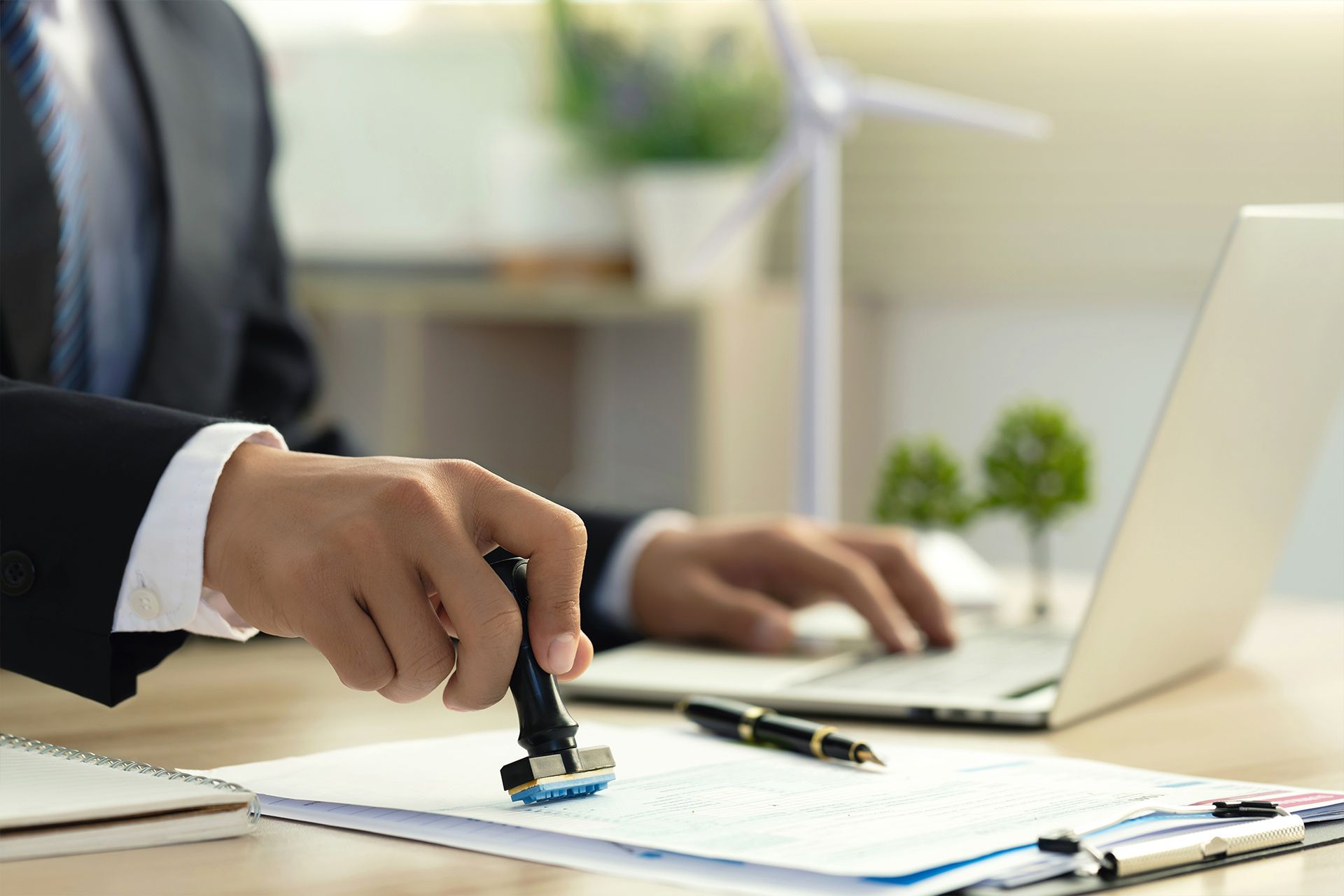 Person in a suit stamping a document with a desk, laptop, and pen in view.