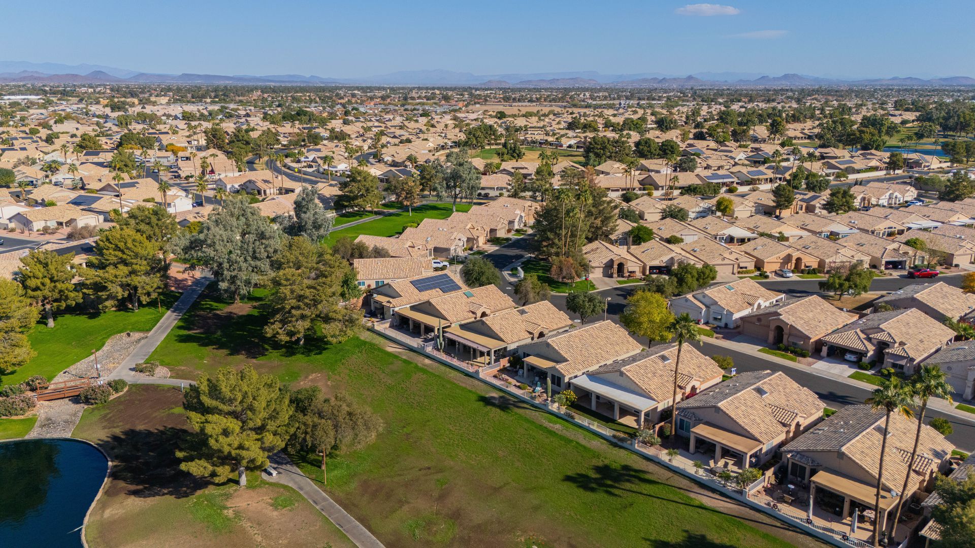 Aerial view of a suburban neighborhood with rows of tan-roofed houses, green parkland, and distant city skyline