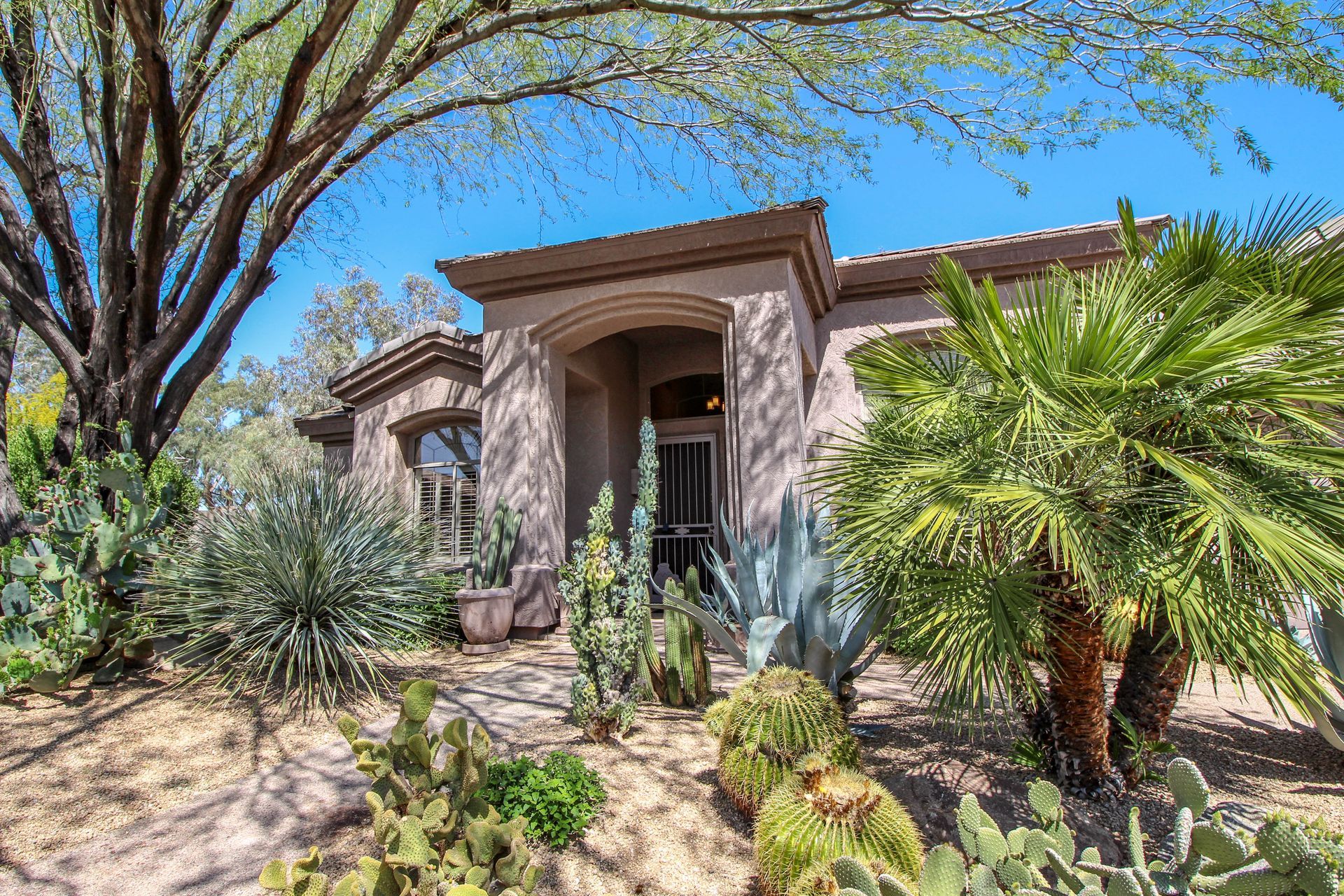 Stucco house entrance with stone archway, desert landscaping, and palm trees under a bright blue sky