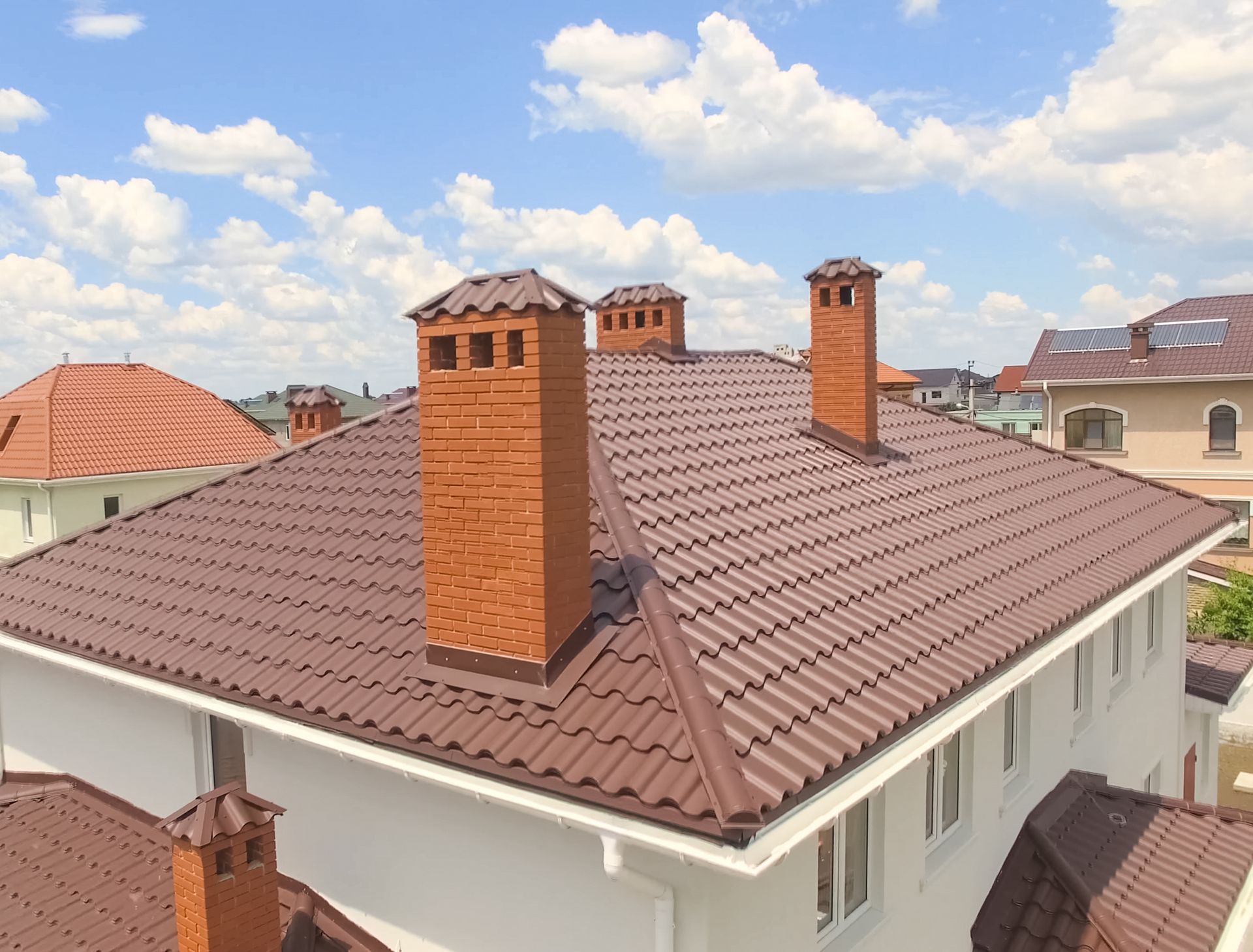 Residential rooftop with brown metal tiles, three brick chimneys, and a cloudy blue sky.