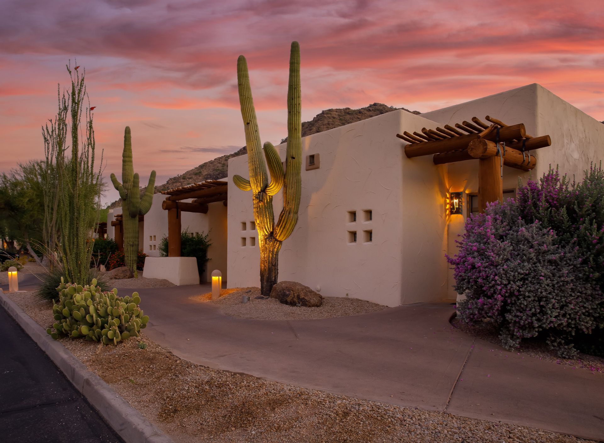 Adobe-style desert home at dusk with cacti, warm lights, and a pink sunset sky