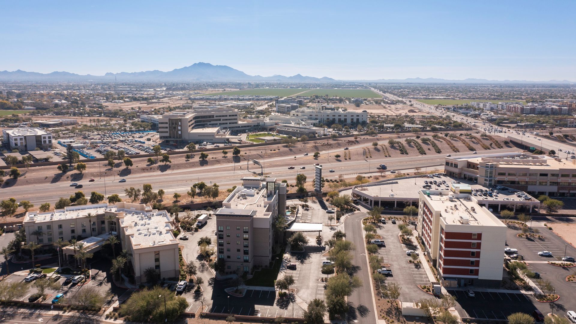 Wide cityscape with low-rise buildings, roads, and a distant mountain under a clear blue sky