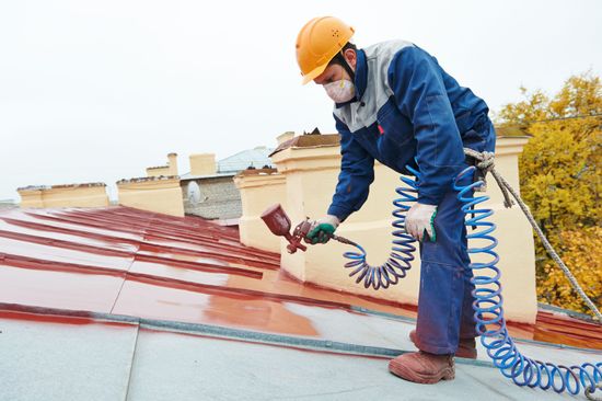 Worker using a spray gun to coat a rooftop with red paint, wearing safety gear and a hard hat Worker using a spray gun to coat a rooftop with red paint, wearing safety gear and a hard hat