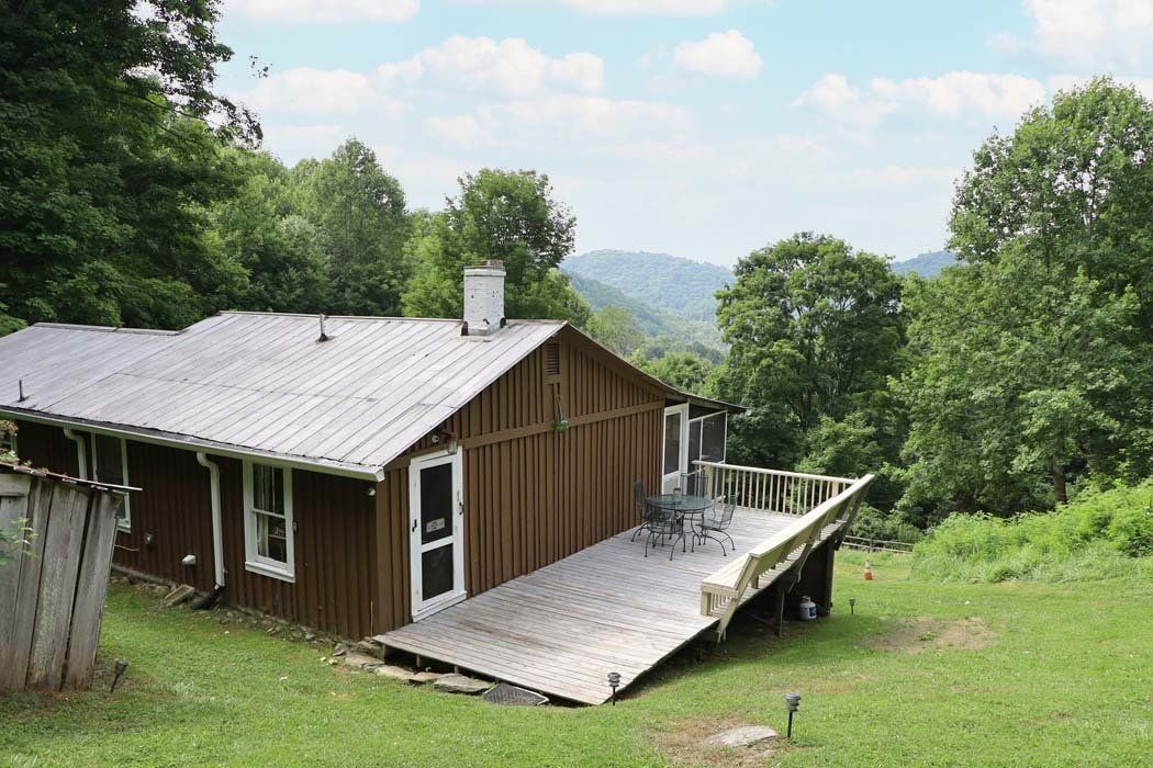 A small house with a large deck in the middle of a grassy field surrounded by trees.