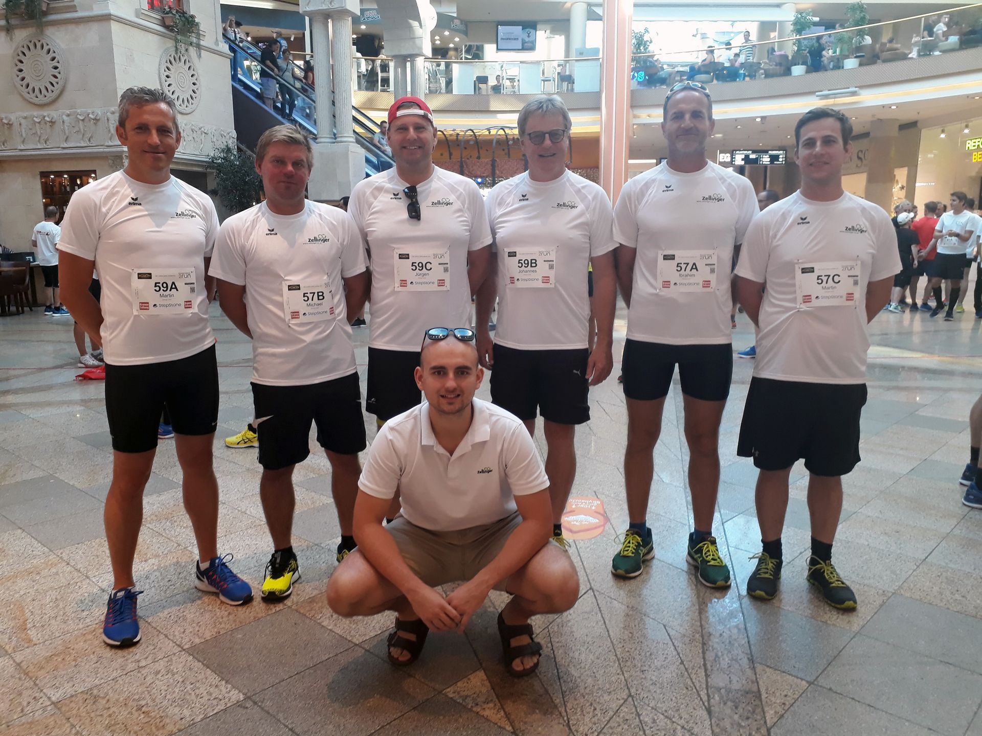 Group of runners in white shirts, black shorts, posed in an indoor setting, one crouched in front.