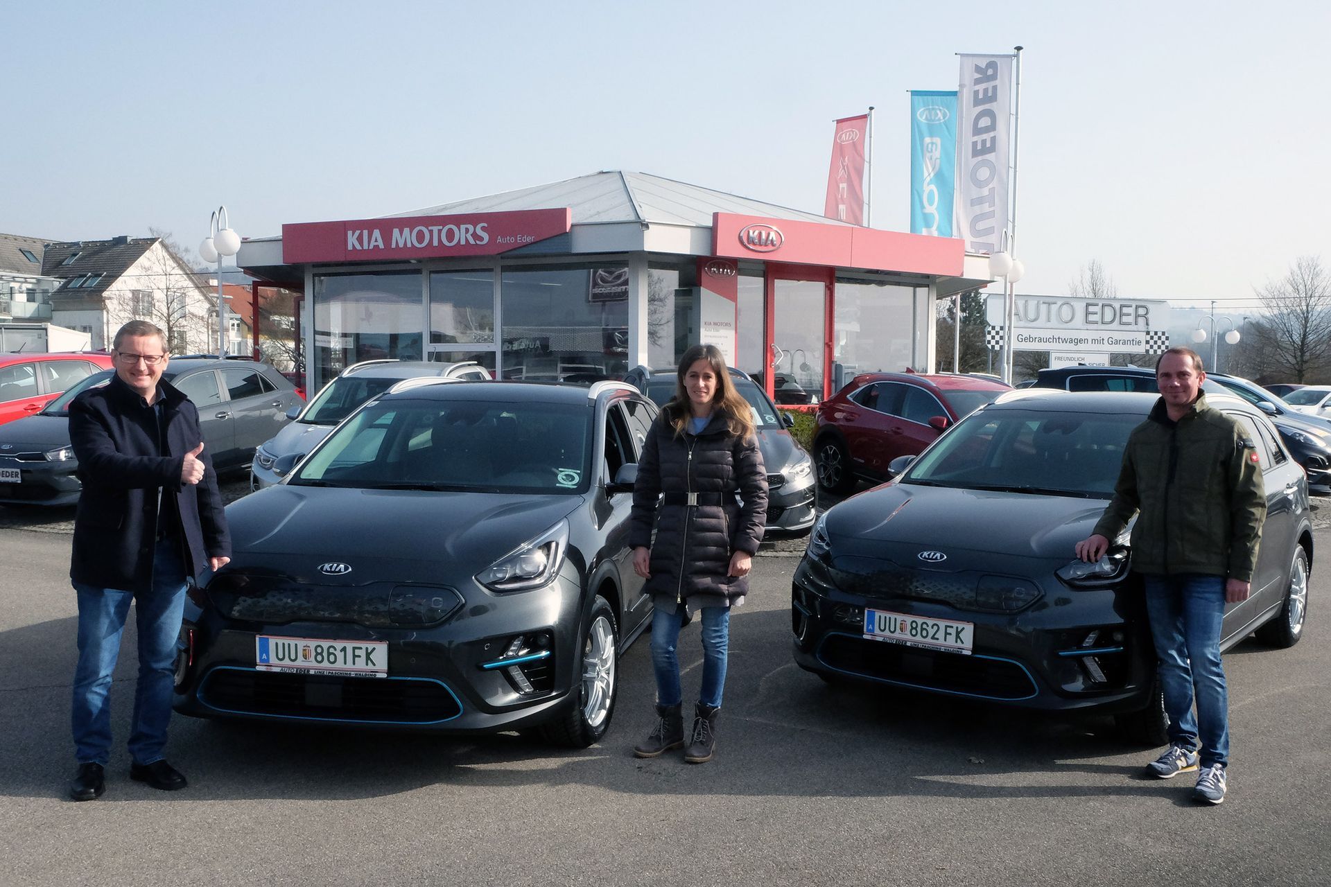 Three people pose with two dark cars at a car dealership. Buildings and flags in the background.