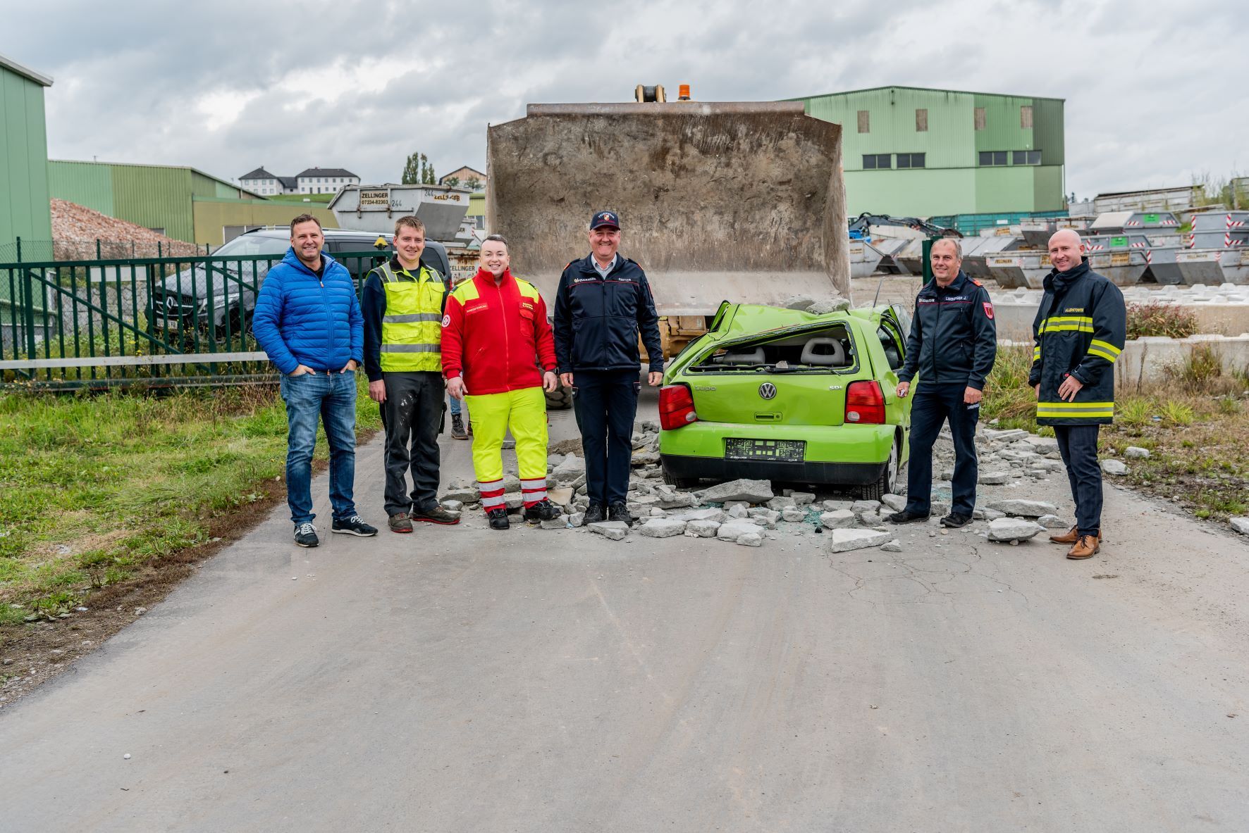 Group of people standing by a crushed green car. A brick wall is behind the car. Concrete debris litters the road.