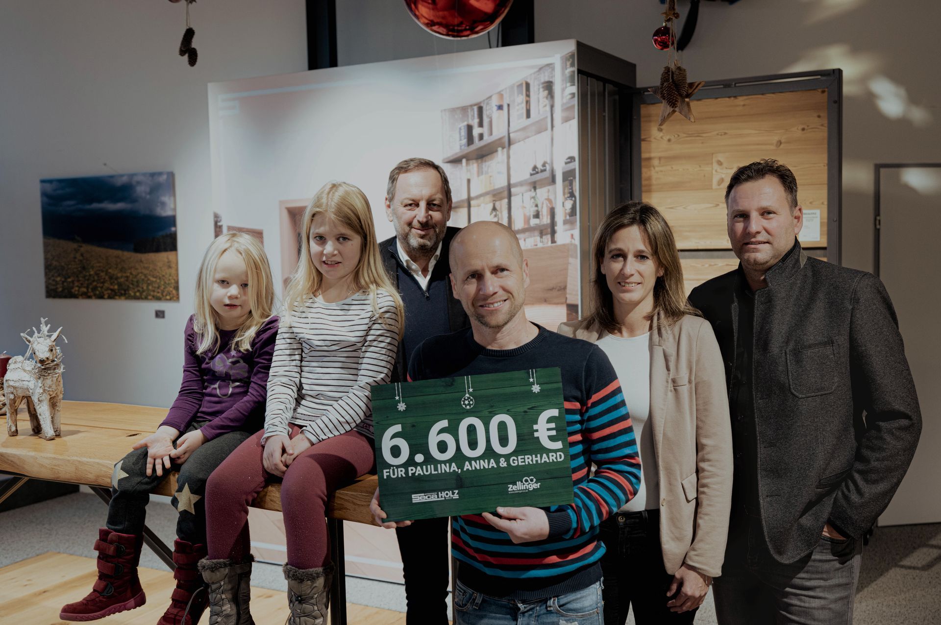 Group holding a check for €6,600. Two young girls sit, people stand, and the setting appears indoors.