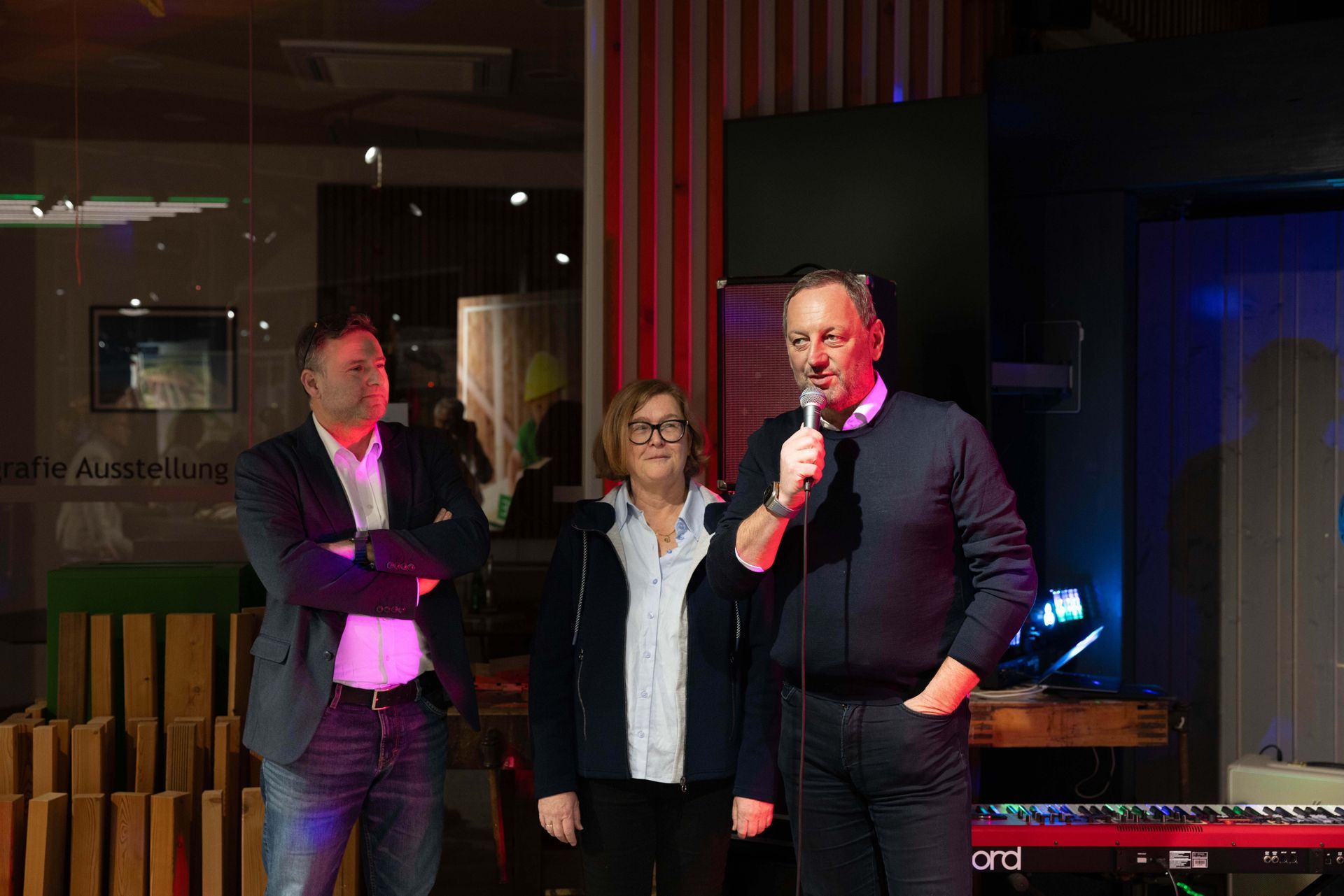 Drei Personen bei einer Veranstaltung. Ein Mann spricht in ein Mikrofon, flankiert von zwei anderen. Die Szene spielt sich in einem Innenraum ab, mit einem Fenster und einer Tastatur.