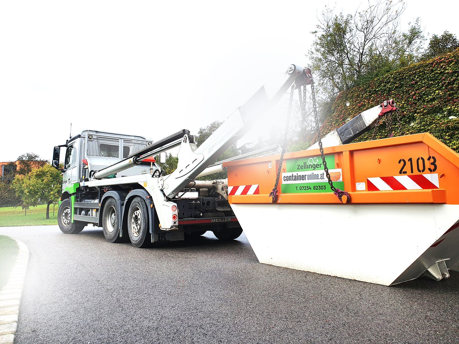 A truck lifting an orange and white dumpster on a wet road.