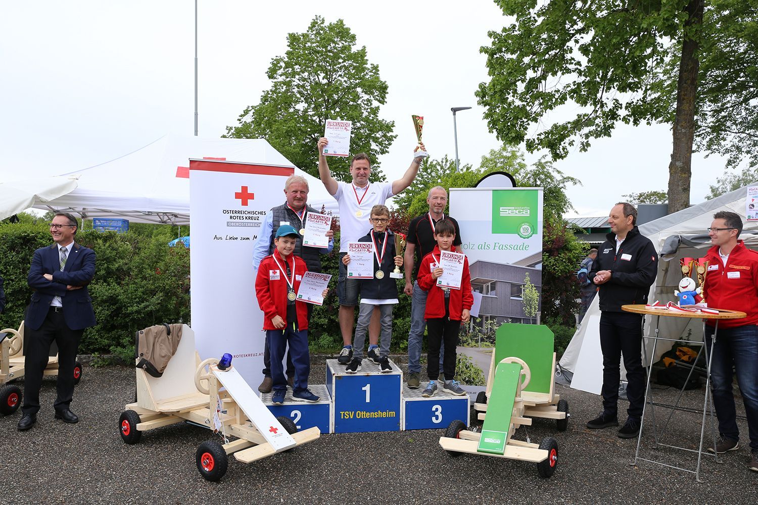 Podium with winners of a Red Cross event, holding trophies and certificates. Outside with carts.