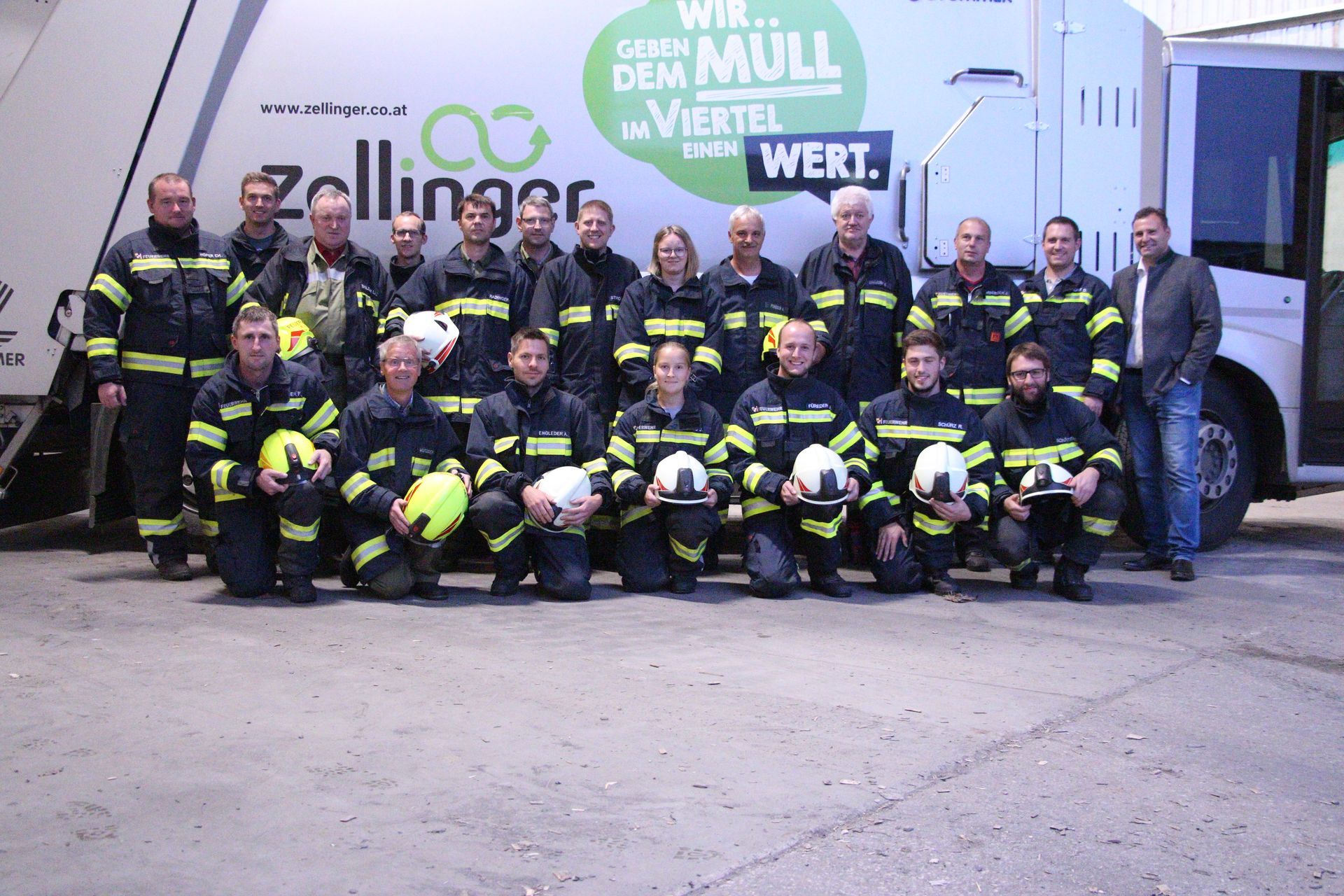 Group of firefighters pose in front of a garbage truck. Many are kneeling, all are wearing yellow stripe jackets.