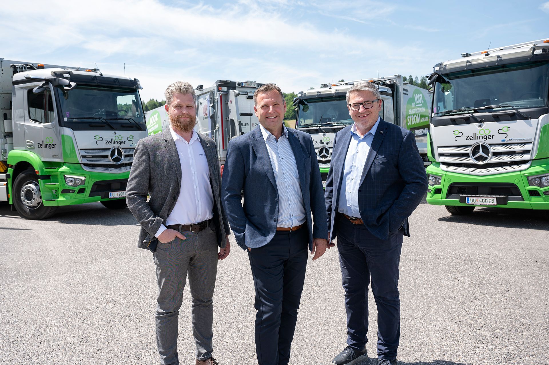 Three men in suits stand in front of green and white trucks. Sunny day.