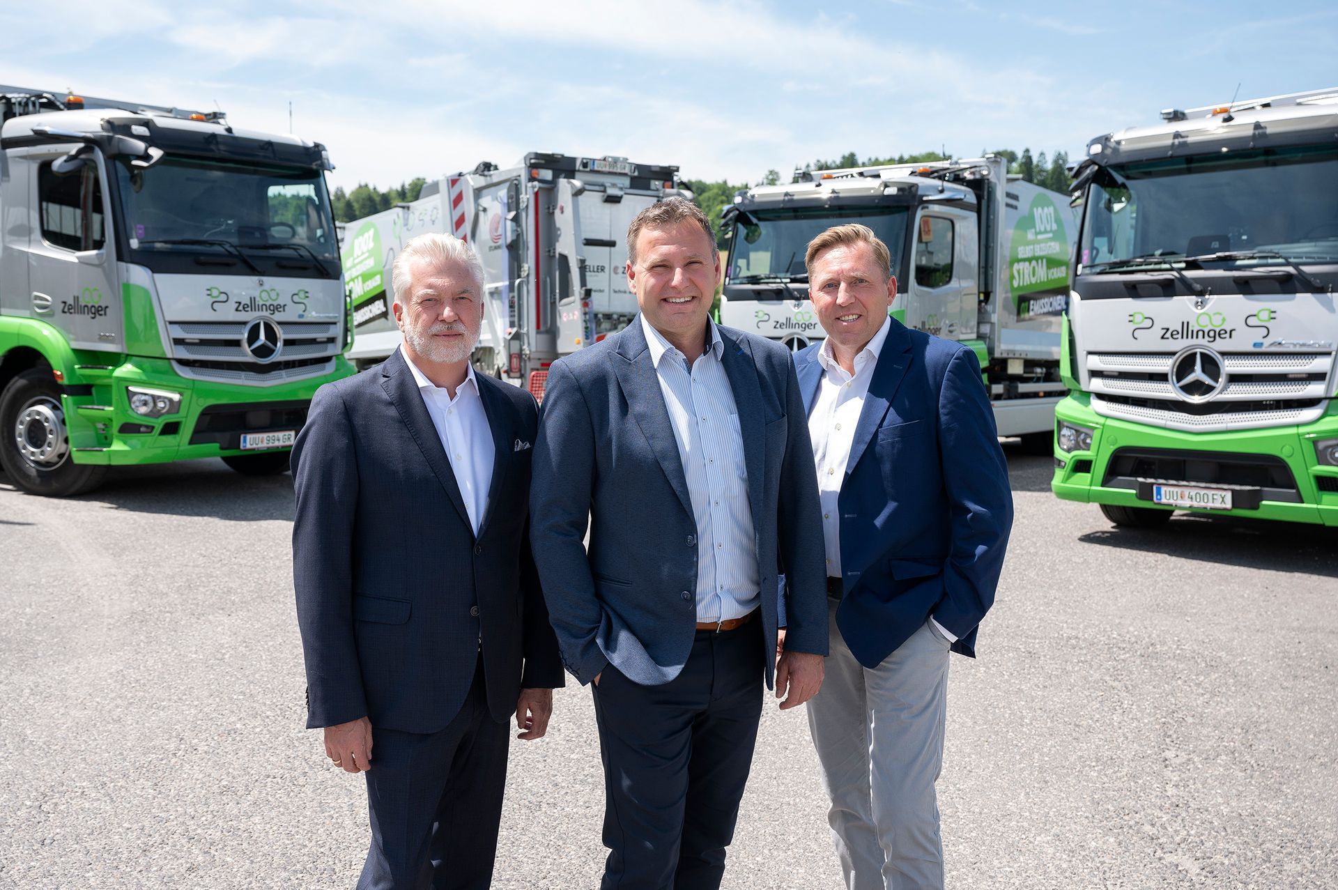 Three men in suits stand in front of several green and silver trucks.