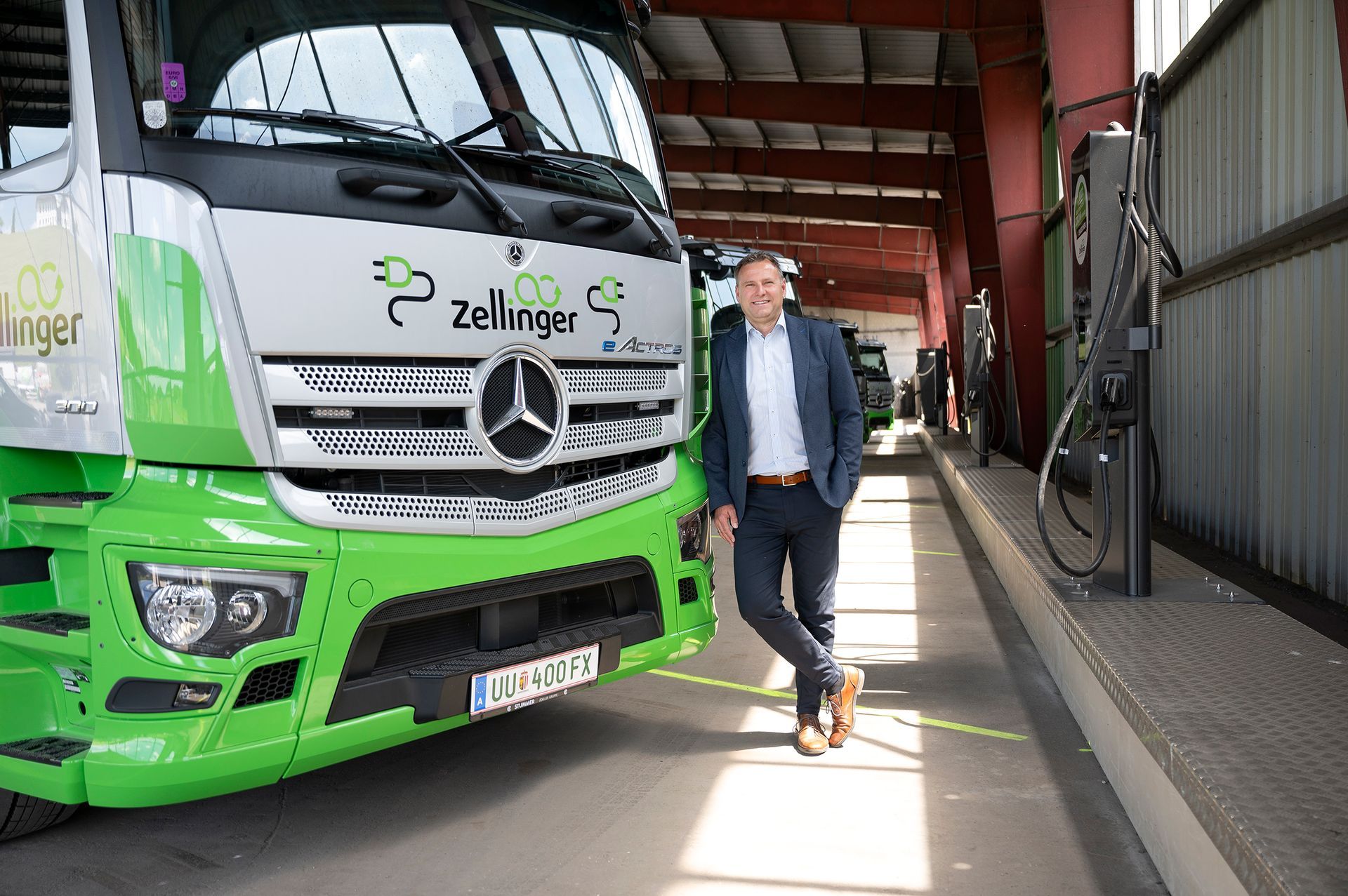 Man in a suit poses by a green and white truck with a logo in a covered area.
