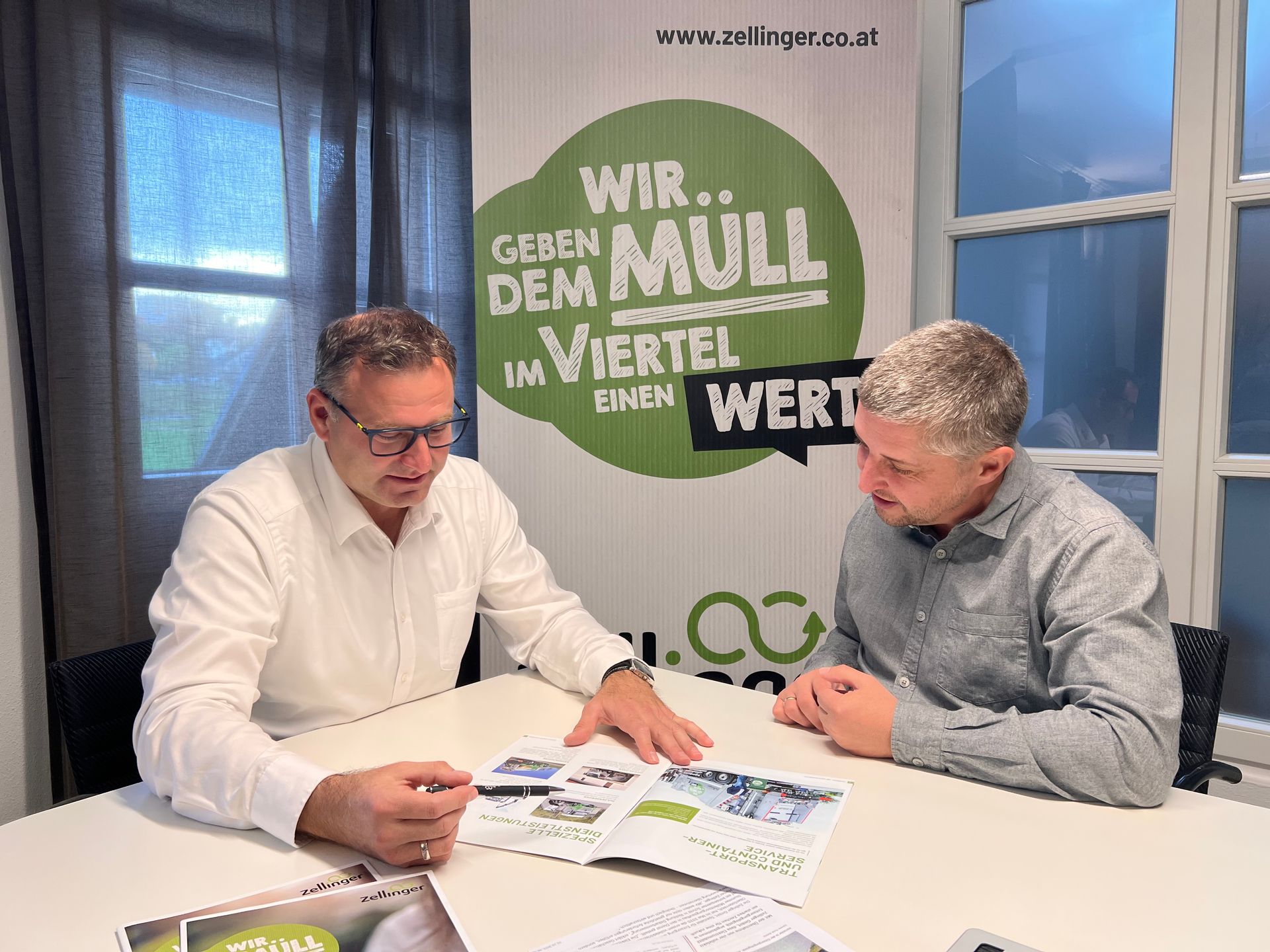 Zwei Männer schauen sich an einem Tisch eine Broschüre an, im Hintergrund ein Banner.