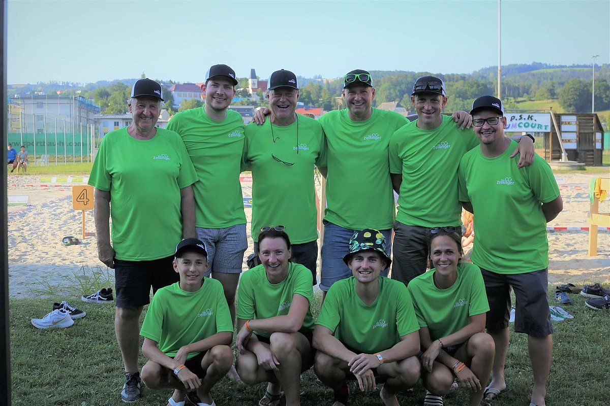 Group of people in green shirts posing on a beach volleyball court. Some are standing, some are kneeling.