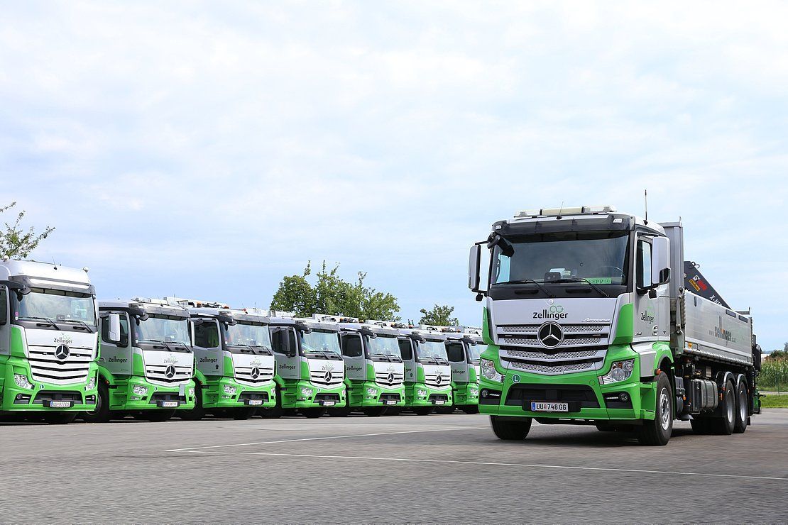 A line of green and silver semi-trucks parked on a paved lot under a cloudy sky.