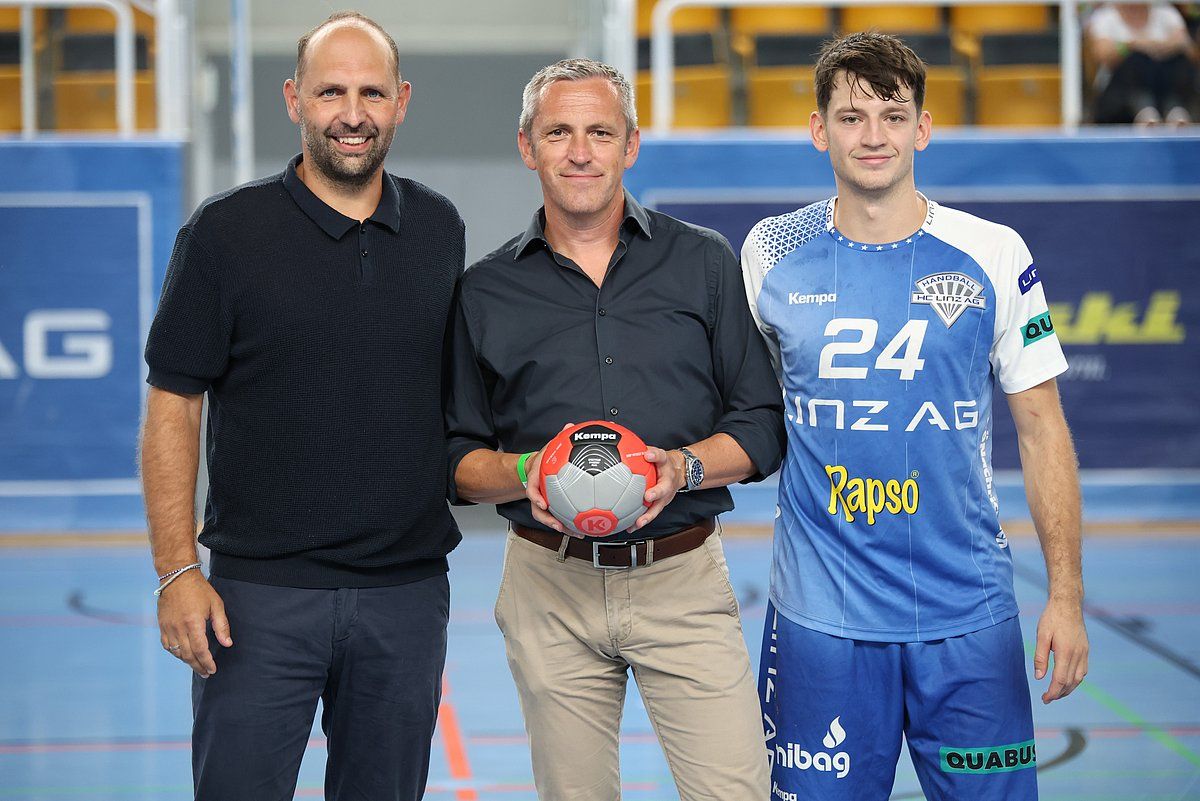 Three men posing with a handball, one in jersey, inside a sports arena.