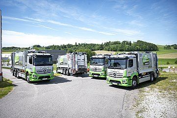 Four green and gray garbage trucks parked on a road with green landscape and blue sky.
