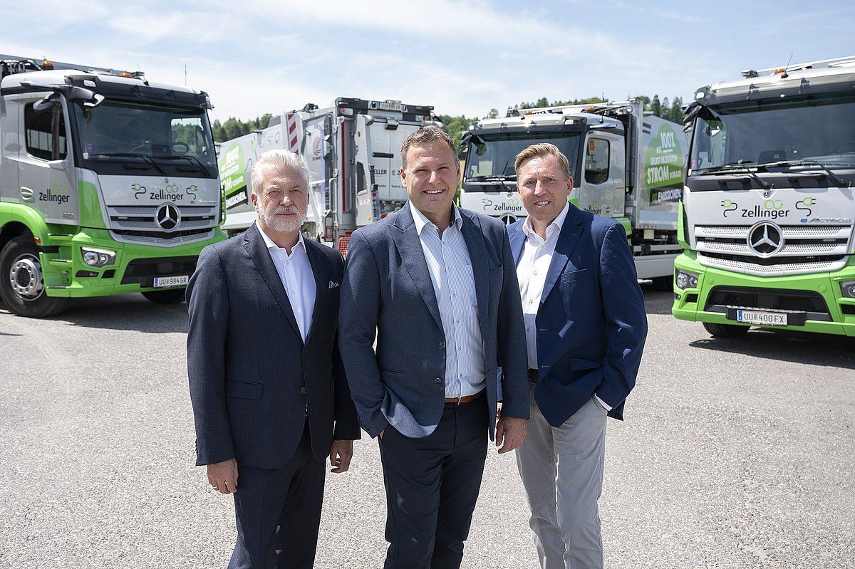 Three men in suits standing in front of a line of trucks with green accents.