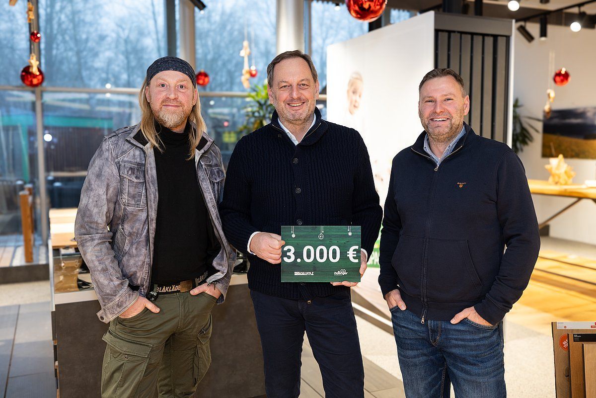 Three men, one holding a check for 3,000 euros, posing indoors. Christmas decorations are visible.