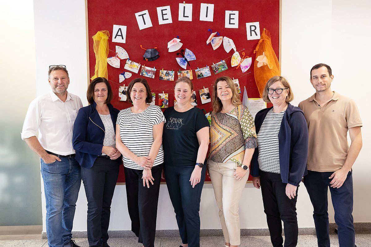 Group of seven people posing in front of a red bulletin board with 