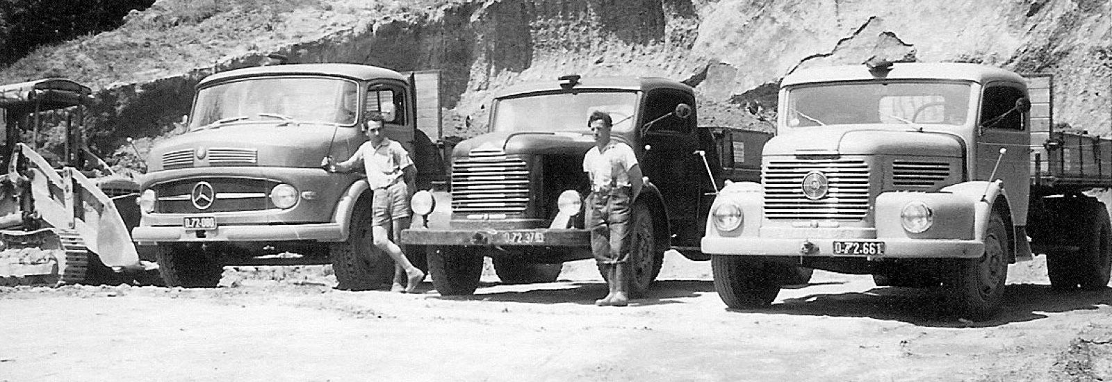 Three vintage trucks parked, with two men standing beside them. Mountainous backdrop. Black and white photo.