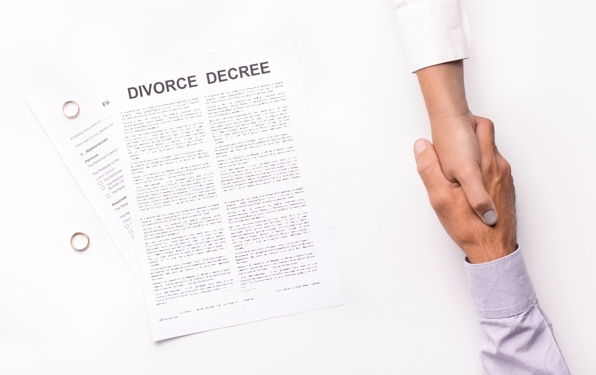 A man’s and woman’s hand are grasping each other amicably, next to papers that read “Divorce Decree. A man’s and woman’s hand are grasping each other amicably, next to papers that read “Divorce Decree.