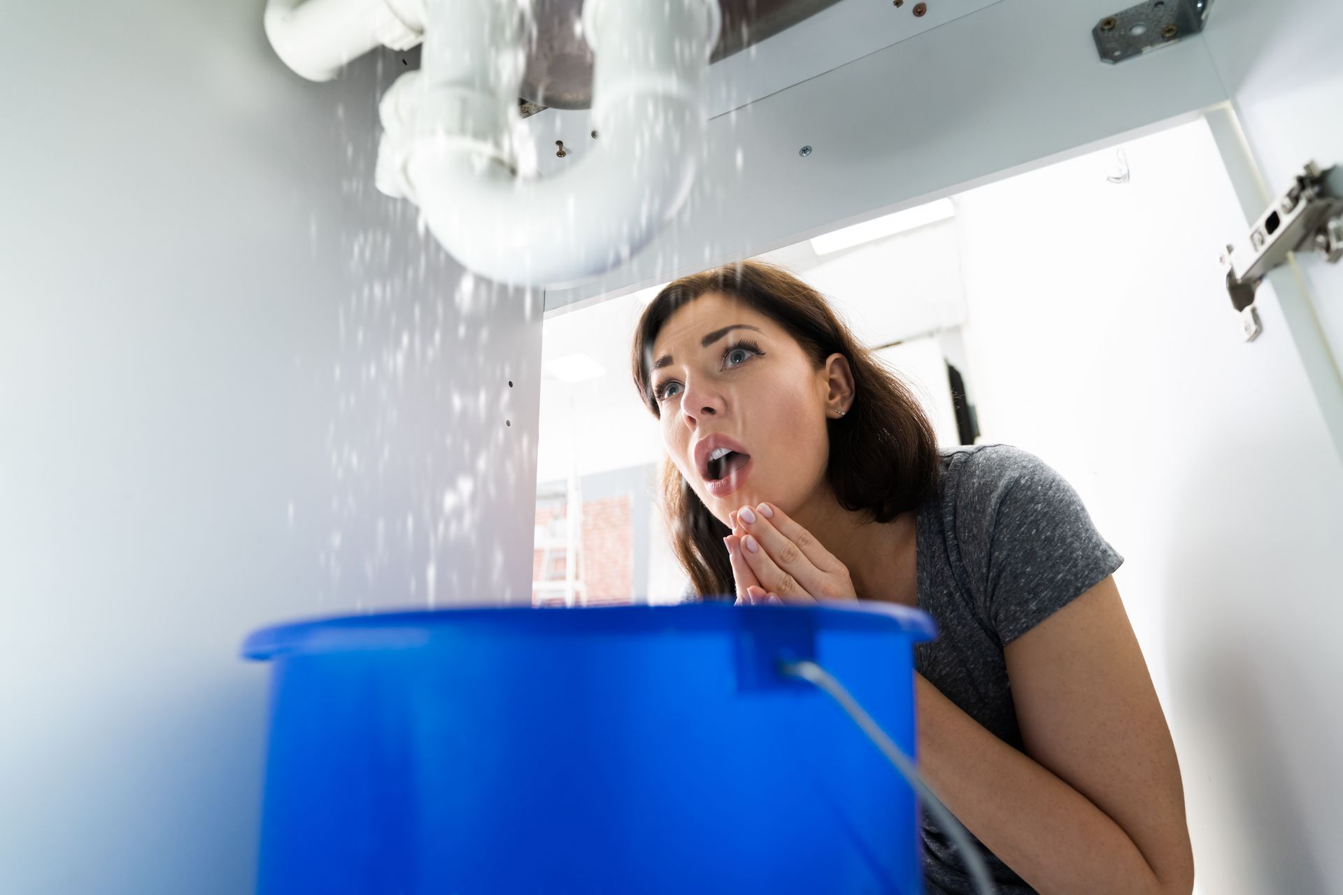Water leaking from under a kitchen sink into a blue bucket during a pipe failure.