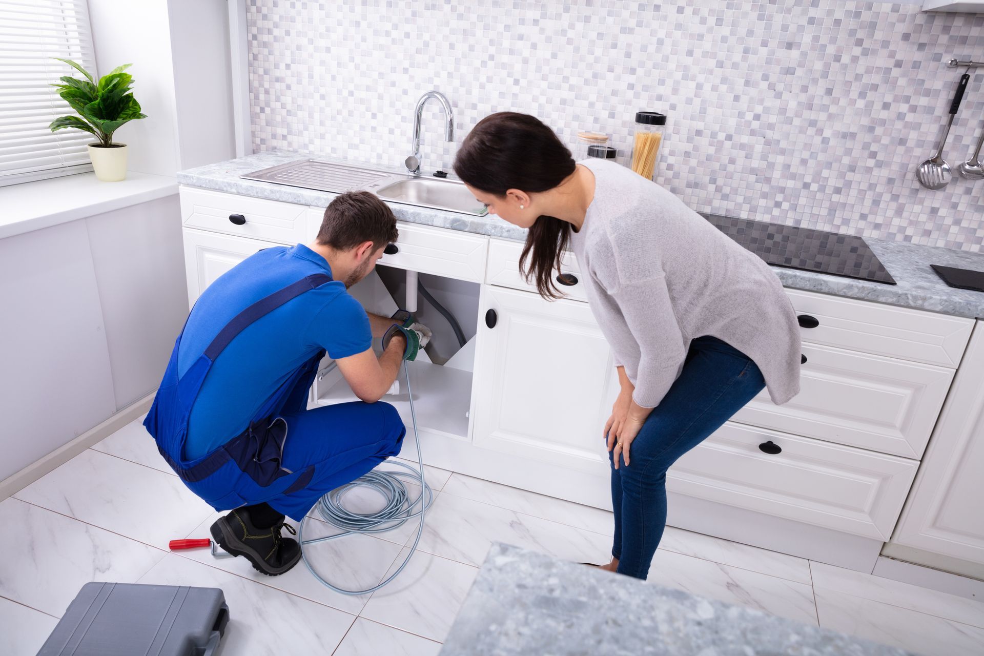 Plumber fixing kitchen sink as woman watches. White cabinets, tiled backsplash, tools and wires visible.
