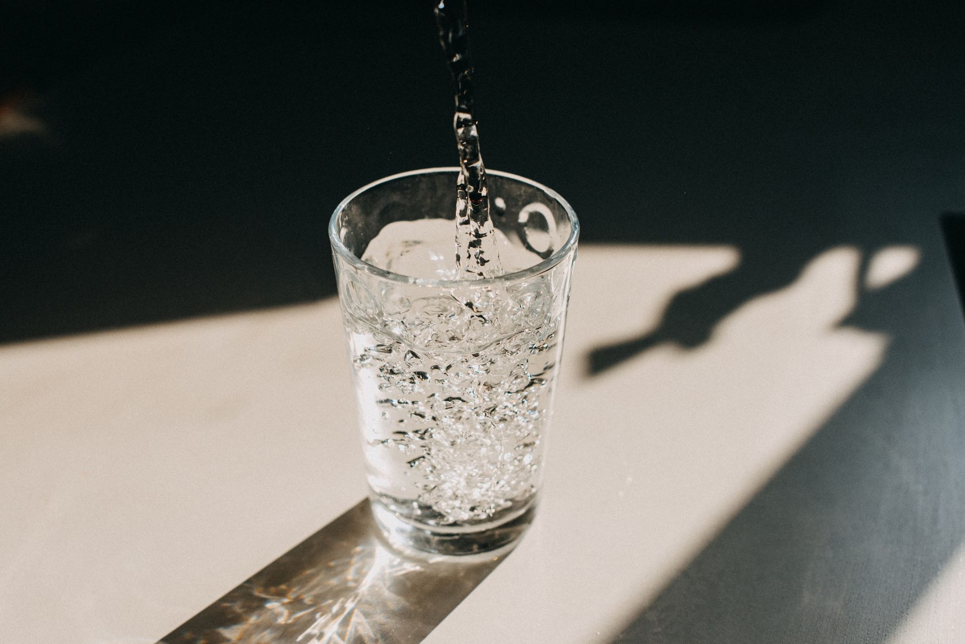 Water pouring into a clear glass, casting shadows on a light-colored surface.