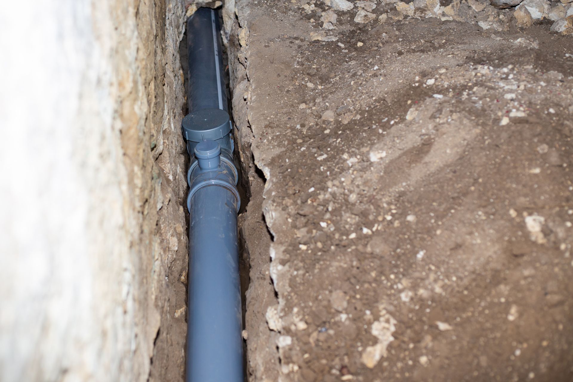 Gray pipe in a trench between a concrete wall and dirt ground.