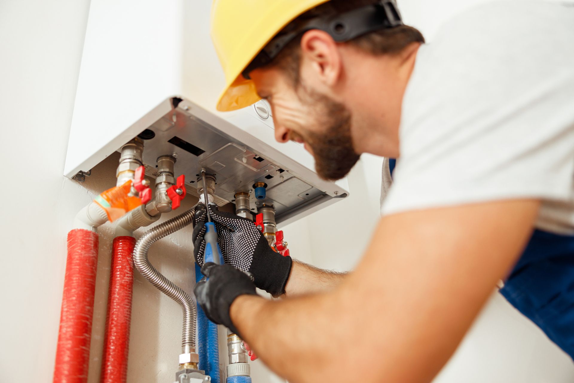 A person wearing a hard hat works on pipes connected to a white wall-mounted appliance.