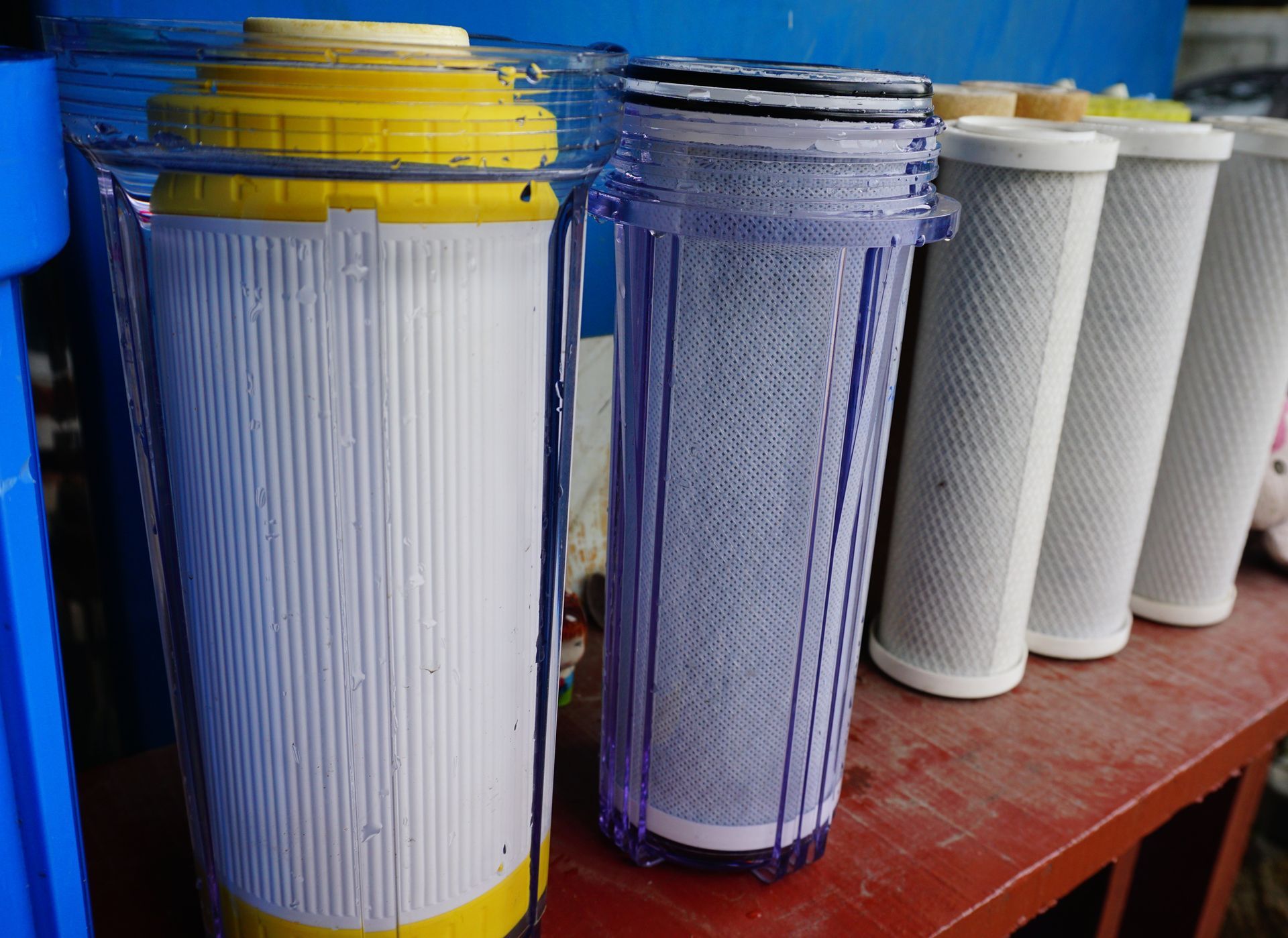 Several water filter cartridges, some encased in transparent housings, lined up on a shelf.