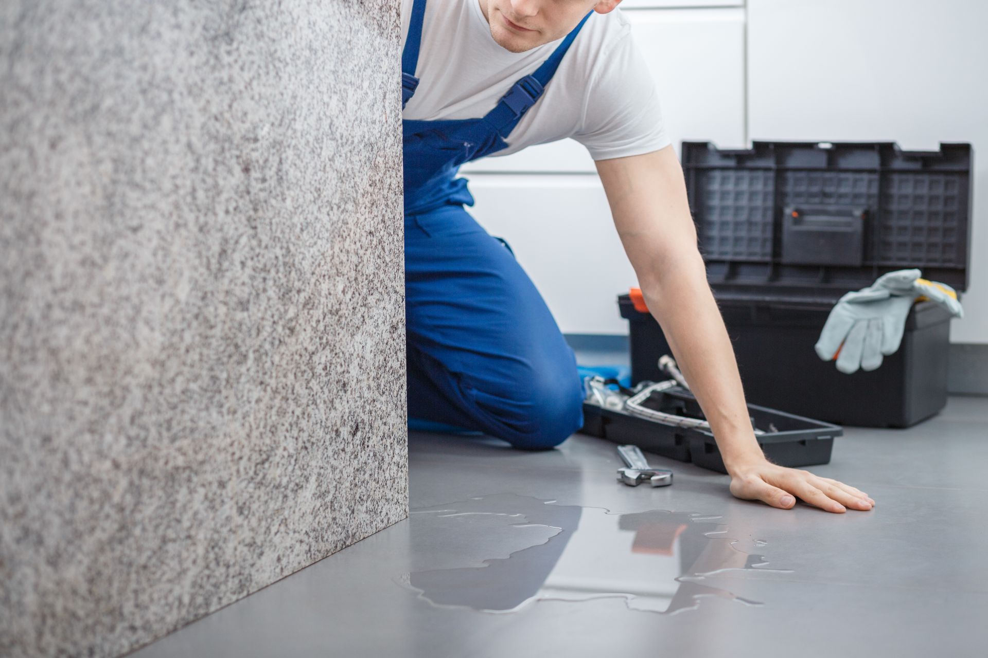 A person in blue overalls kneels next to a water leak on a gray floor, with tools in a toolbox.