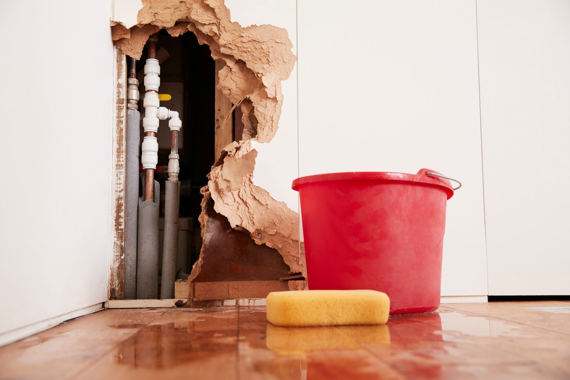 Wall damaged with exposed pipes. Red bucket and yellow sponge on wet floor.