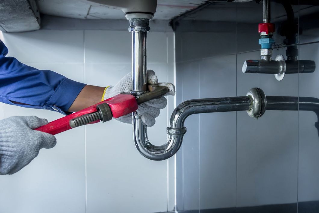 Plumber in blue shirt and gloves using a wrench on a sink drain pipe.