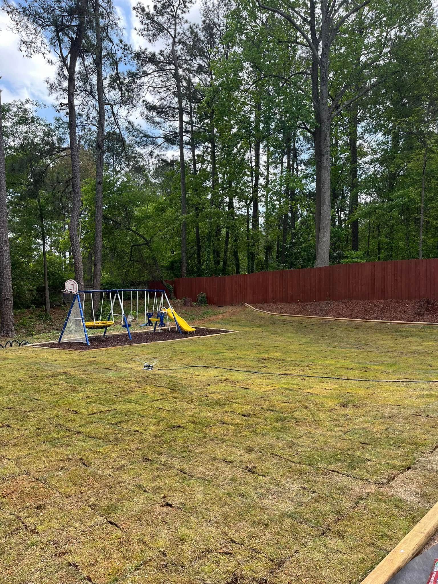 There is a playground in the backyard with a red fence and trees in the background.