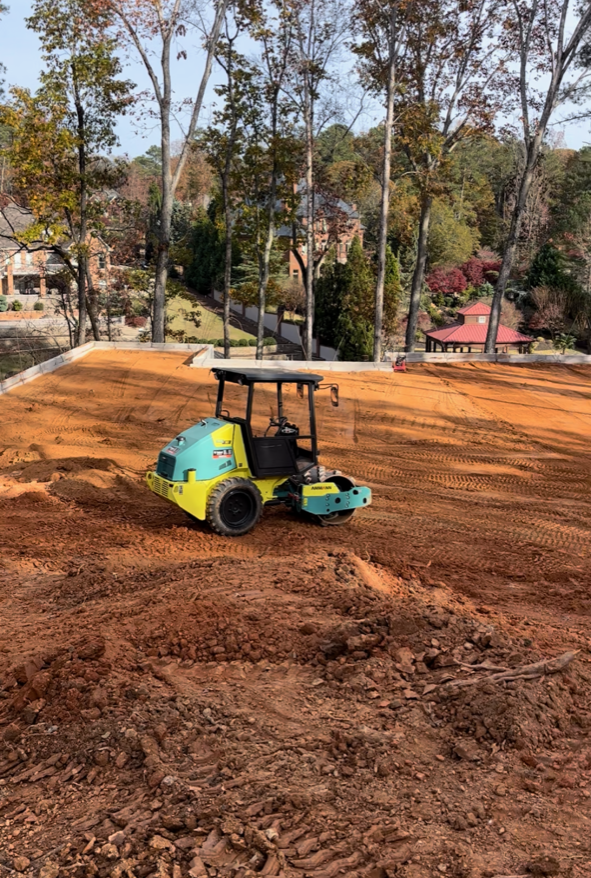 A blue and yellow roller is moving dirt in a field.